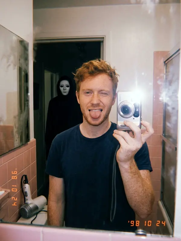 Man taking a mirror selfie with a masked figure behind him