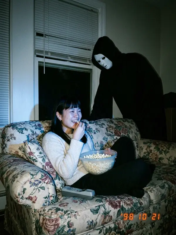 Woman on a floral couch with popcorn, masked figure leaning in